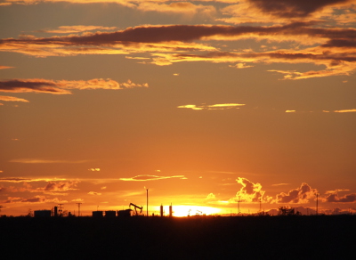 [A close-in view of the horizon where it appears the setting sun has set the ground on fire. The bright-white color stretches across a section of the horizon backlighting an oil derrick, three storage tanks, and several columns. This color also outlines all the clouds low in the sky. The ones higher up are also lit, but not as brightly.]